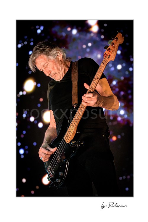 Roger Waters leans forward while playing a black bass with a natural wood neck, wearing a black shirt against scattered colorful background lights.