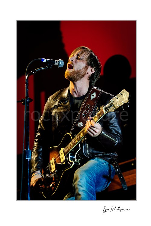 Dan Auerbach of The Black Keys Live Concert Print  Vertical color image of Dan Auerbach of The Black Keys singing into a microphone while playing guitar against a vivid red stage backdrop