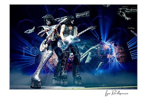 Horizontal color photograph of Paul Stanley and Tommy Thayer performing side by side in full makeup and costumes with bright stage lighting, hexagonal screens, and pyrotechnics behind them