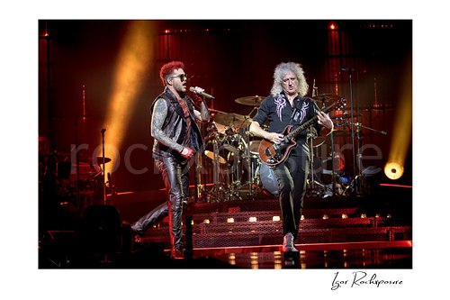 Horizontal color image of Adam Lambert and Brian May of Queen performing onstage together with the drummer visible behind them under warm red concert lighting