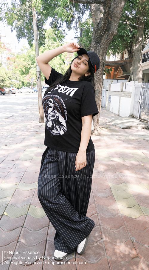 Nupur Ahuja wearing a black KR$NA Rapper T-shirt, black cap, and pinstripe trousers while posing playfully on a tree-lined street for Nupur Essence urban fashion photography