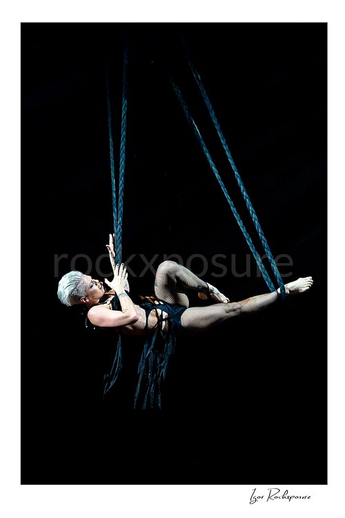 Vertical color image of Pink suspended high above the stage on ropes in a dramatic aerial live performance pose