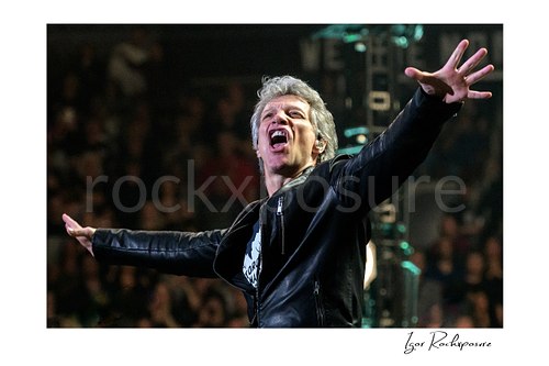 Horizontal color image of Jon Bon Jovi in a dramatic live performance close-up, singing with both arms outstretched and the arena crowd blurred behind him