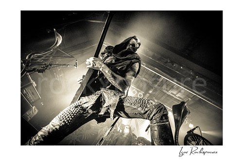 Horizontal black and white image of Nergal of Behemoth performing with a mic stand in a dramatic low-angle stage shot under intense backlight