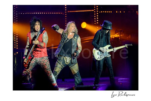 Horizontal color image of Nikki Sixx, Vince Neil and Mick Mars performing live on stage during a Mötley Crüe concert with bass guitar, microphone and warm concert lighting