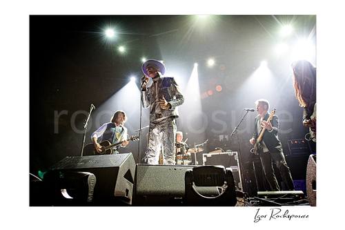 Horizontal color image of The Tragically Hip performing live on stage, with Gord Downie in a glittering silver suit at center under bright concert spotlights and band members visible across the stage