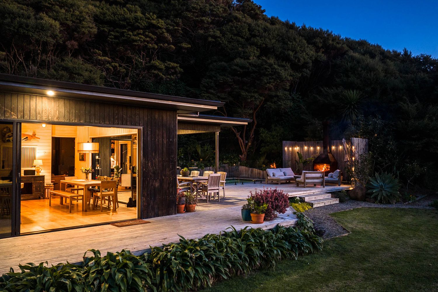 Twilight architectural photography of a luxury modern home in Raglan showing an illuminated indoor living area, outdoor patio, and bush backdrop, captured by Flax Cove Studio