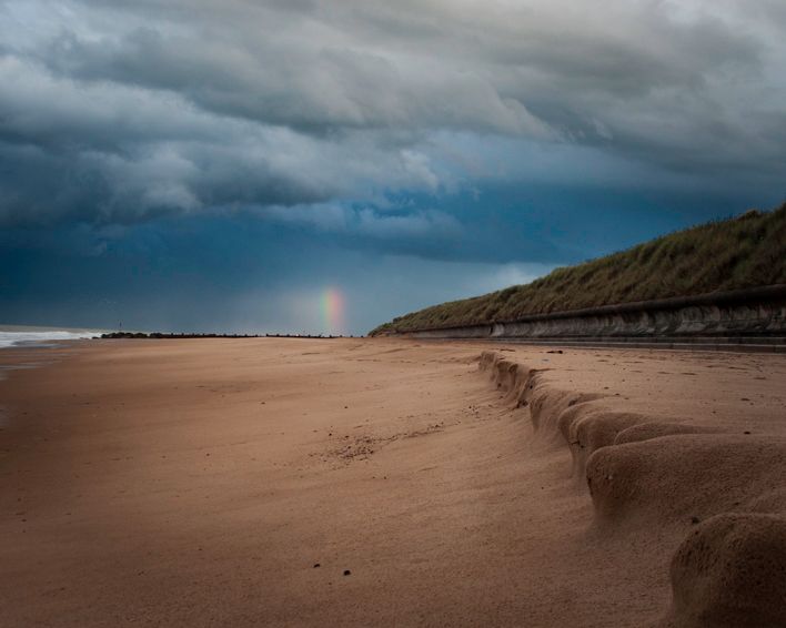 A distant rainbow breaking through heavy dark storm clouds over a wide sandy beach and eroded dunes on the Norfolk coast.