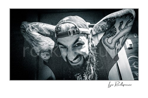 Horizontal black and white image of Mike Portnoy grinning at the camera with tattooed arms raised behind his head and a backward cap