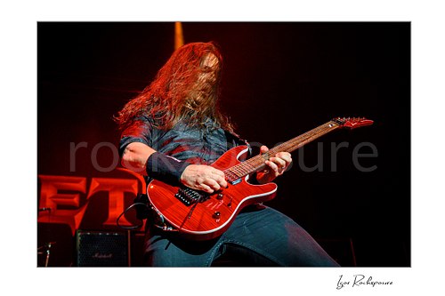 Horizontal color image of Kiko Loureiro of Megadeth playing guitar on stage under red lighting with long hair across his face