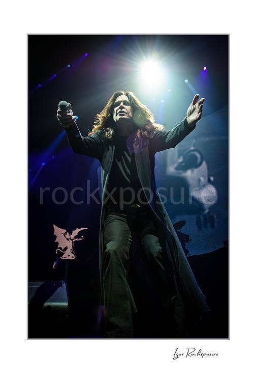 Ozzy Osbourne of Black Sabbath standing with both arms raised on stage under dramatic blue concert lighting during a live performance