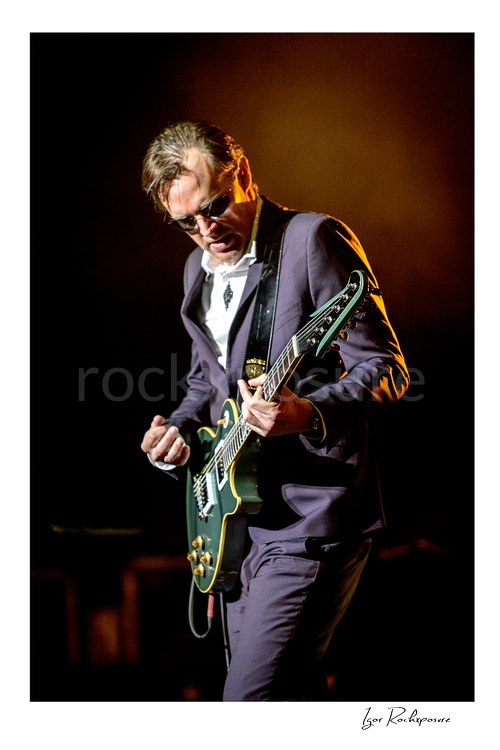 Vertical color image of Joe Bonamassa performing live on stage with his guitar under warm stage lighting