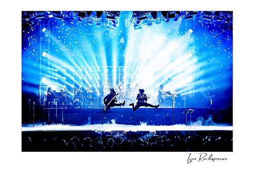Horizontal color image of Trans Siberian Orchestra performing on stage with two jumping guitarists, bright blue light beams, and a packed concert crowd