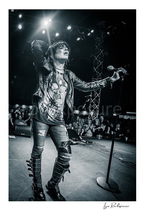 Vertical black and white image of Tom Keifer performing live on stage with one hand behind his head and the other on a microphone stand, with the crowd visible behind him