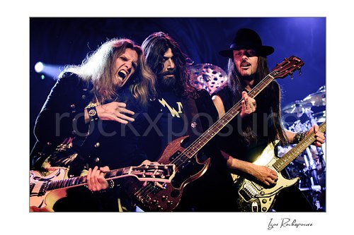 Horizontal color image of Joel Hoekstra, Michael Devin, and Reb Beach of Whitesnake performing live on stage, with Hoekstra at left, Devin at center on bass, and Beach at right playing guitar under vivid blue and purple concert lighting  Whitesnake members Joel Hoekstra