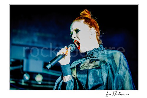 Horizontal color image of Shirley Manson singing into a microphone during a Garbage concert in a dramatic close-up under cool blue stage light