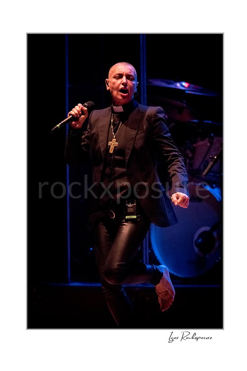 Sinéad O’Connor sings holding a microphone with one bare foot lifted behind her, wearing a black jacket as blue light illuminates the drum kit.