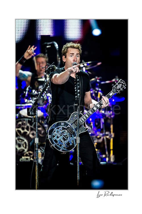 Vertical color image of Chad Kroeger of Nickelback singing into a microphone and pointing toward the crowd while playing guitar with the drummer visible behind him