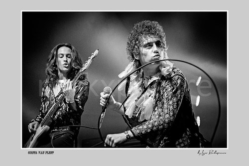 Horizontal black and white image of Greta Van Fleet members Sam Kiszka on the left playing bass guitar and Josh Kiszka on the right holding a microphone on stage