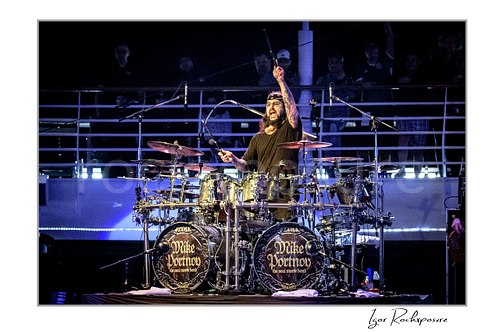 Horizontal color image of Mike Portnoy playing at a double bass drum kit with one drumstick raised high under blue stage lighting