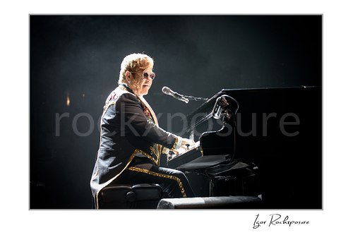 Horizontal color image of Elton John playing piano under a bright white spotlight in a dark concert scene