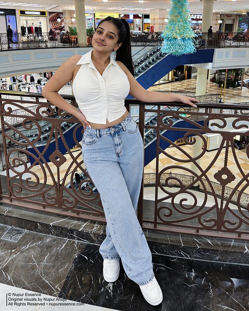 Nupur Ahuja posing at a shopping mall balcony in a white halter top and light wash denim jeans, featured through Nupur Essence