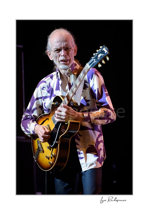 Vertical color image of Steve Howe of Yes playing guitar onstage in a patterned shirt during a live performance
