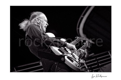 Horizontal black and white image of Billy Sheehan playing his double-neck bass in an energetic side-stage performance shot