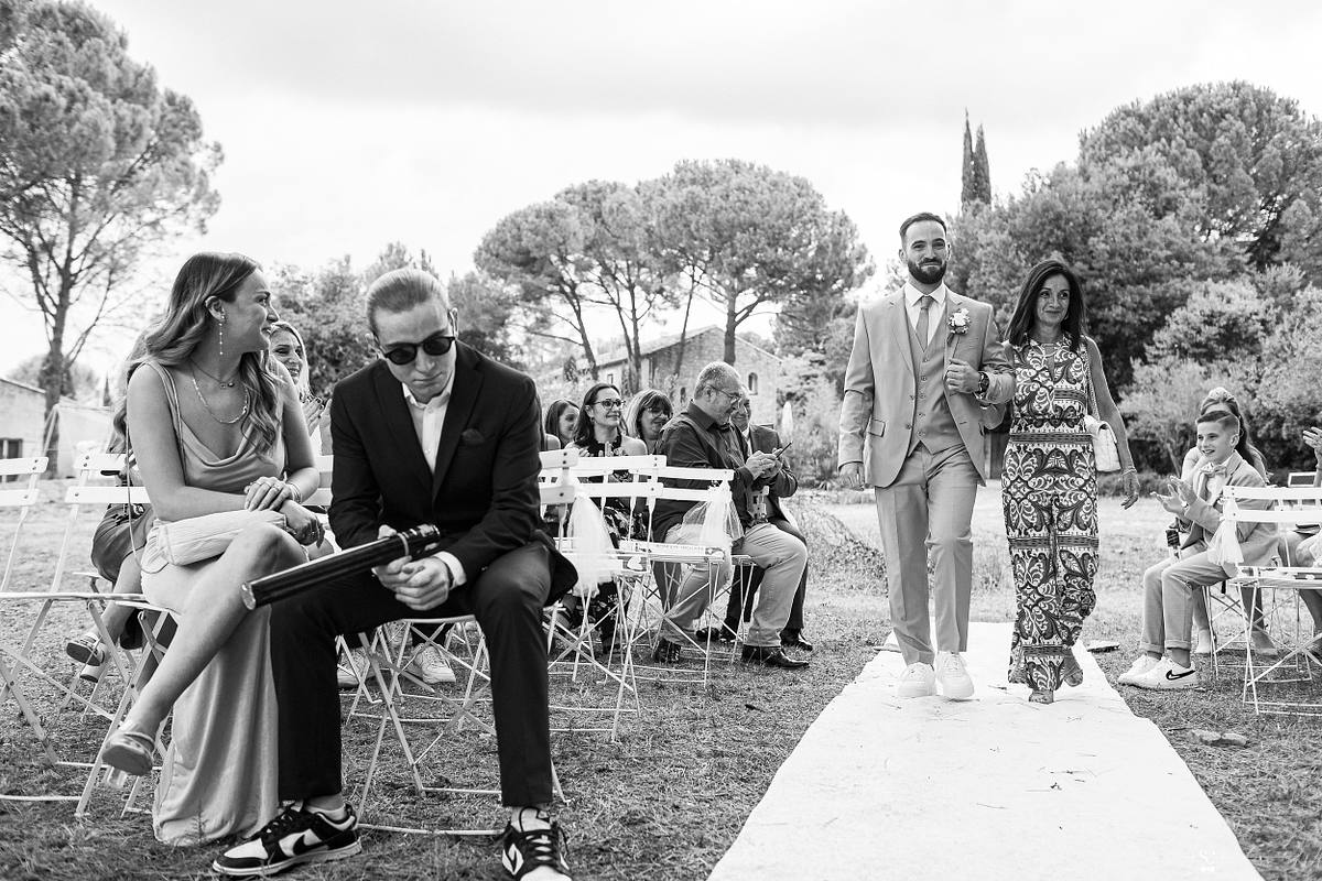 Mari&eacute; qui rentre aux bras de sa maman marchent ensemble sur un tapis blanc, entour&eacute;s de leurs invit&eacute;s sous les arbres de la prairie, capturant un moment de bonheur partag&eacute; et l'ambiance d&eacute;contract&eacute;e du mariage c&eacute;l&eacute;br&eacute; &agrave; N&icirc;mes