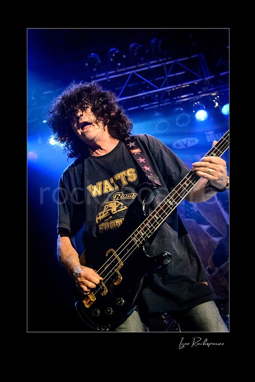 Vertical color image of Jimmy Bain playing bass onstage under blue concert lighting in an energetic live performance shot