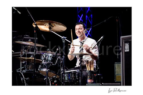 Horizontal color image of Laust Sonne smiling at the drum kit during a live performance, with drumsticks raised and stage lighting visible