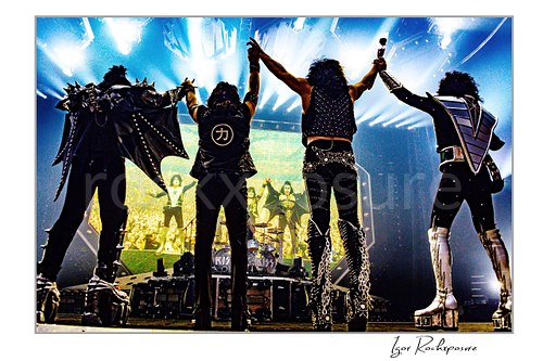 Horizontal color photograph of the KISS band standing onstage with their backs to the camera holding hands in the air while facing a massive stage screen that displays their own image, creating a dramatic mirrored moment in front of the crowd