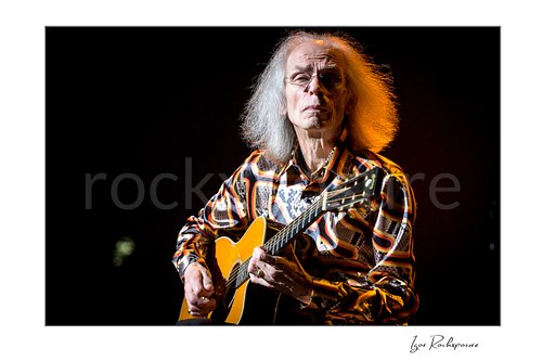 Horizontal color image of Steve Howe of Yes playing acoustic guitar onstage under warm concert light