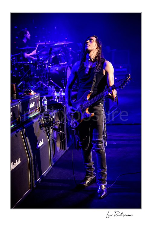 Vertical color image of Nuno Bettencourt standing on stage with an electric guitar under deep blue lighting, with Marshall amplifiers beside him