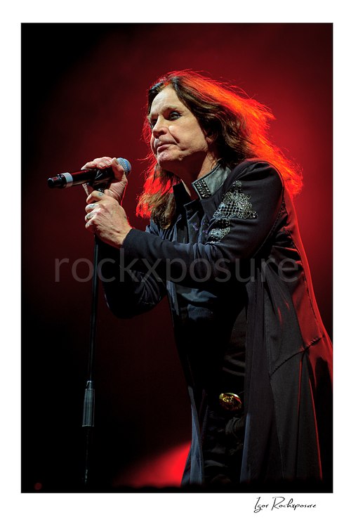 Vertical concert photography of Ozzy Osbourne of Black Sabbath singing into a microphone under dramatic red stage lighting