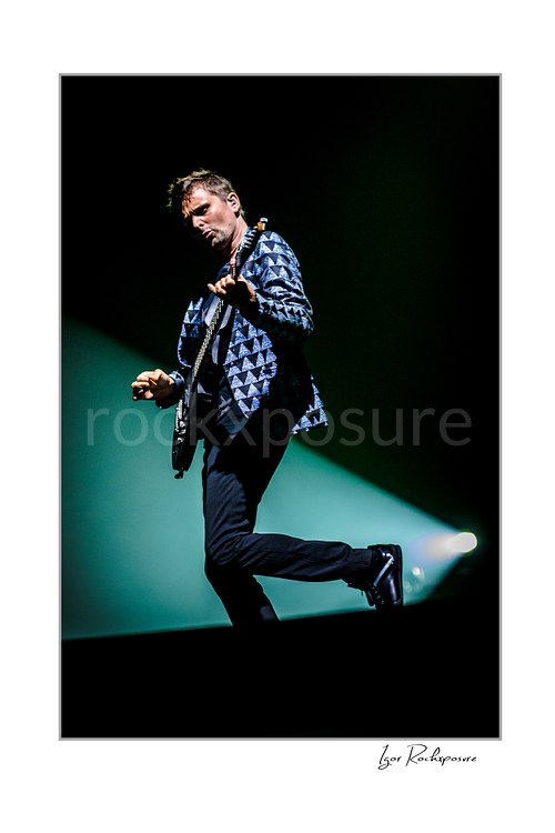 Vertical color image of Matt Bellamy of Muse playing guitar in a patterned stage jacket with one leg lifted under dramatic spotlight