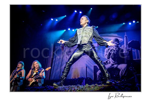Horizontal color image of Steve Harris, Dave Murray, and Bruce Dickinson of Iron Maiden performing live on stage, with Harris at left, Murray next to him on guitar, and Dickinson in a dramatic wide stance at center under blue concert lights.