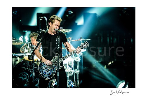Horizontal color image of Chad Kroeger of Nickelback singing into a microphone while playing guitar with the drummer visible behind him under bright concert beams
