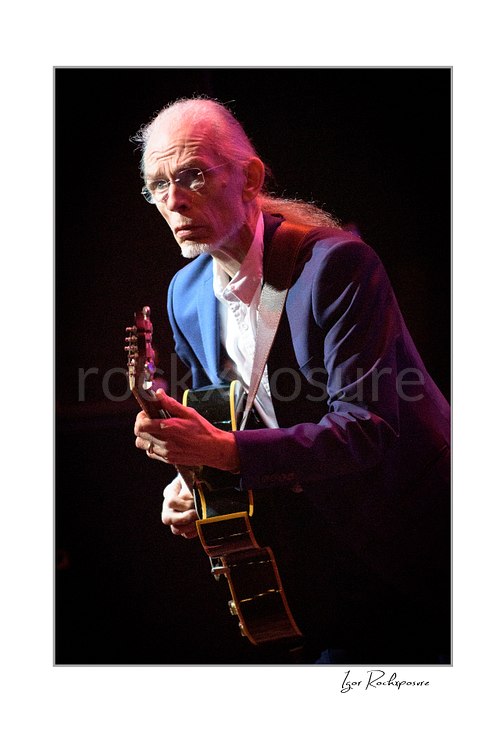 Vertical color image of Steve Howe of Yes playing guitar onstage under warm red concert light