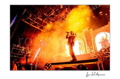 Horizontal color image of M. Shadows of Avenged Sevenfold singing into a handheld microphone on stage with dramatic orange light, fire, and stage smoke