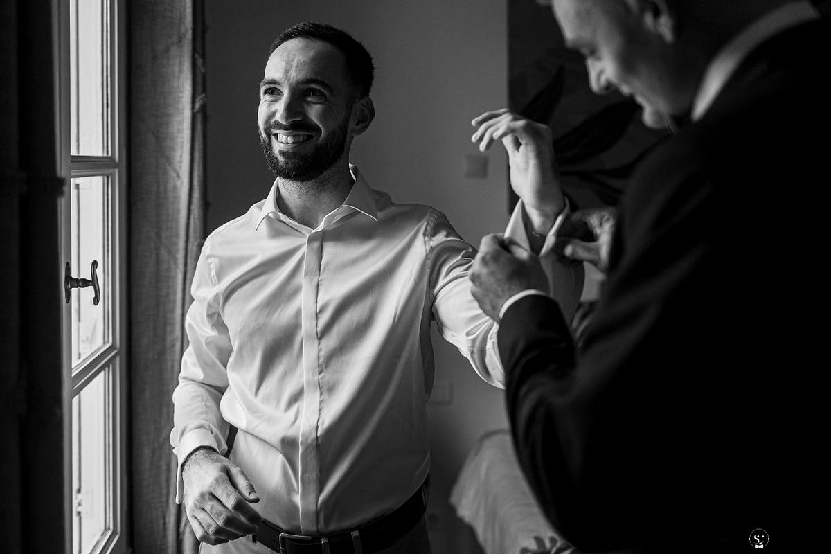 Un instantan&eacute; joyeux du mari&eacute; en chemise blanche souriant largement, &eacute;clair&eacute; par la lumi&egrave;re naturelle d'une fen&ecirc;tre, avec son t&eacute;moin ajustant sa manche, capturant la complicit&eacute; et l'&eacute;l&eacute;gance avant la c&eacute;l&eacute;bration du mariage &agrave; N&icirc;mes