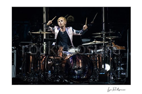 Horizontal color image of Dominic Howard of Muse at the drum kit with both drumsticks raised in a dramatic live performance