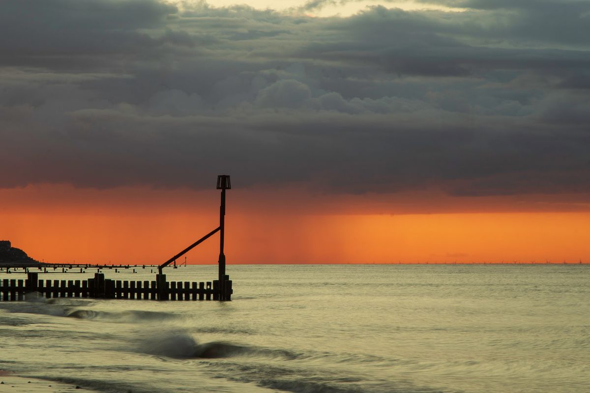 A vibrant orange evening sunset over the North Sea at Walcott beach with a distant rain shower and wooden sea groynes