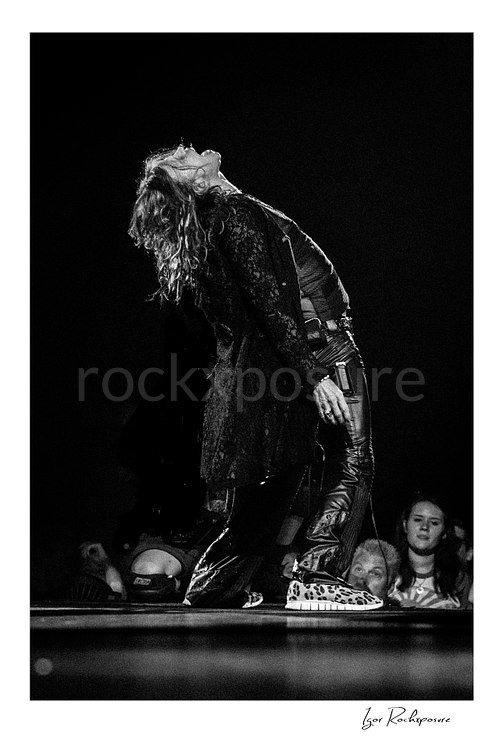Vertical black and white image of Steven Tyler of Aerosmith performing live on stage in a backward-leaning pose with long hair, dark layered clothing, and patterned shoes