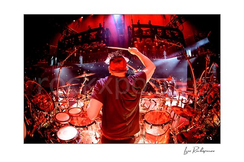 Horizontal color image of Korn drummer Ray Luzier seen from behind at the drums with a drumstick raised overhead under vivid red stage lighting