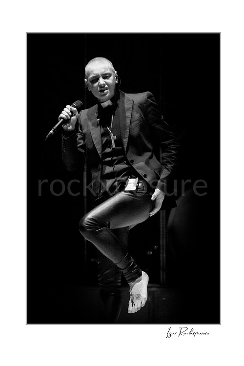 Sinéad O’Connor lifts one knee and bare foot while holding a microphone near her shoulder, wearing a black jacket in a vertical black and white portrait.