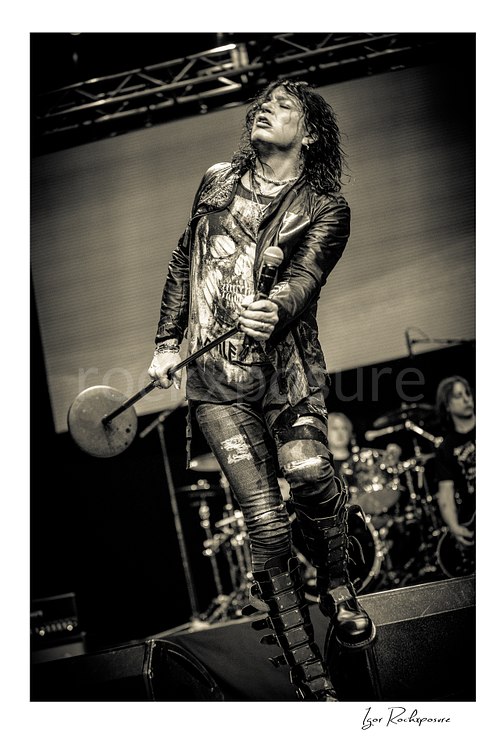 Vertical black and white image of Tom Keifer striding on stage in a dramatic live performance pose, holding a microphone stand with the drum kit visible behind him