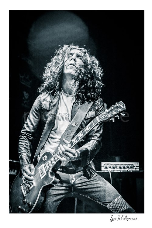 Vertical black and white image of Brent Woods playing guitar on stage with curly hair and dramatic overhead light