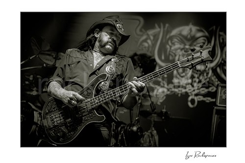 Horizontal black and white image of Lemmy Kilmister of Motörhead performing live on stage, wearing a cowboy hat and playing bass guitar with the band backdrop visible behind him