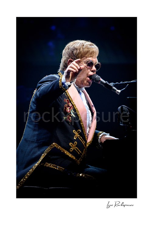 Vertical color image of Elton John singing at the piano with one hand raised under dark blue stage lighting
